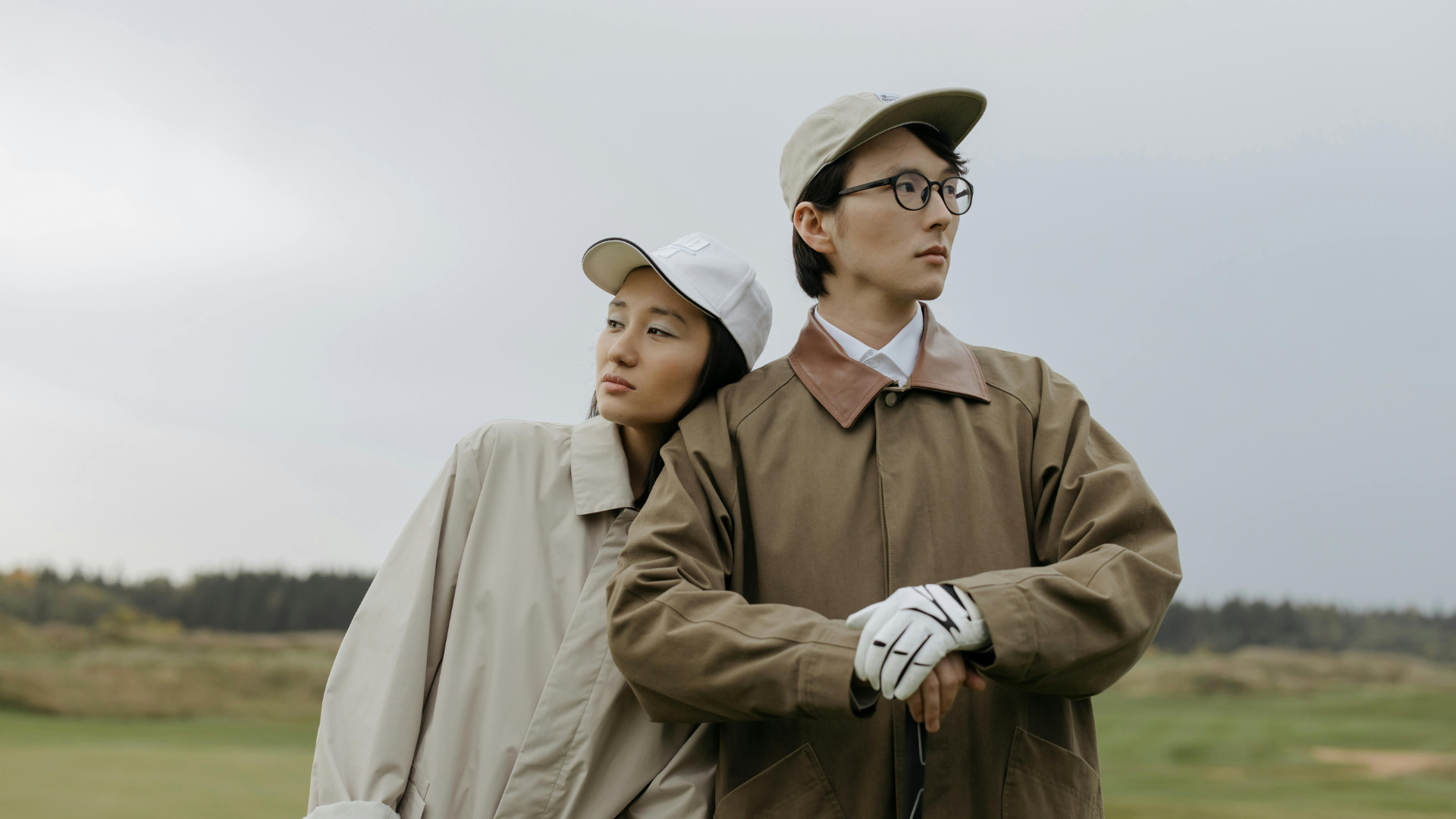 Two people in outdoor clothing standing on a grassy field with a neutral background